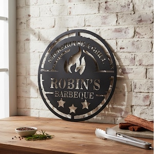 May include: Black metal sign with the text "Smokehouse & Grill" and "Robin's Barbeque" cut out. The sign features a flame design and three stars. It is mounted on a white brick wall.