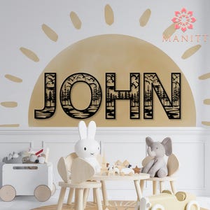 Pode incluir: Arte de parede de madeira preta com a palavra "JOHN" e um design com tema de natureza. As letras apresentam silhuetas de &aacute;rvores, p&aacute;ssaros e &aacute;gua. A arte &eacute; montada em uma parede bege com um design semelhante ao sol, criando um tema decorativo para o quarto das crian&ccedil;as.