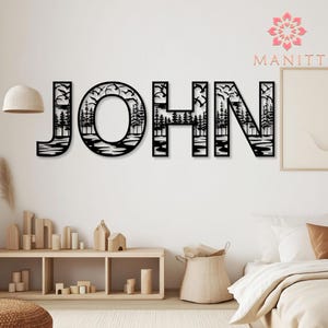 Pode incluir: Arte de parede em metal preto que soletra "JOHN", com uma cena da natureza dentro de cada letra. O design inclui &aacute;rvores, &aacute;gua e p&aacute;ssaros voando, criando uma silhueta c&ecirc;nica. A marca "MANITT" &eacute; exibida.