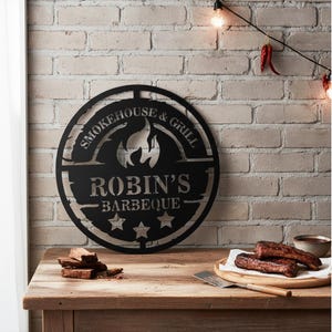 May include: Black metal sign with the text "Smokehouse & Grill" and "Robin's Barbeque" with a flame graphic. The sign is round and has three stars at the bottom. The sign is on a wooden table with food and a brick wall background.