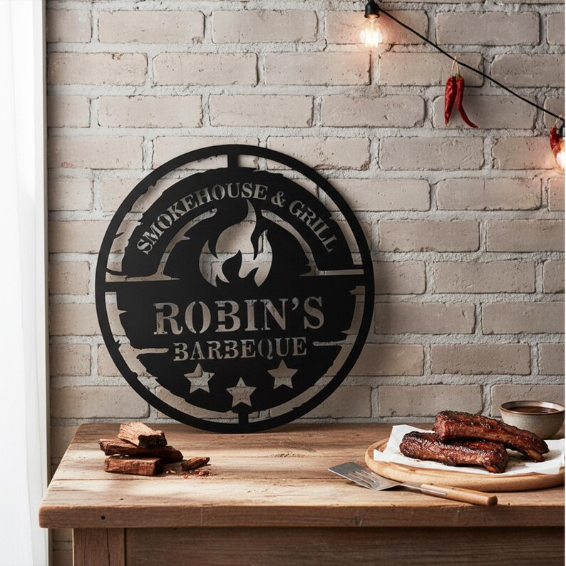 May include: Black metal sign with the text "Smokehouse & Grill" and "Robin's Barbeque" with a flame graphic. The sign is round and has three stars at the bottom. The sign is on a wooden table with food and a brick wall background.