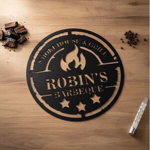 May include: Black metal sign with the text "Smokehouse & Grill" and "Robin's Barbeque" with three stars. The sign has a flame design in the center. The sign is on a wooden surface with wood chips, peppercorns, and a thermometer.