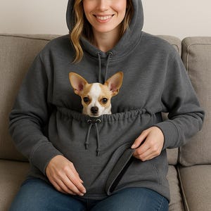 May include: A dark gray hoodie with a kangaroo pouch, featuring a small tan and white dog. The hoodie has a drawstring hood and is worn with blue jeans. The dog is looking directly at the camera.