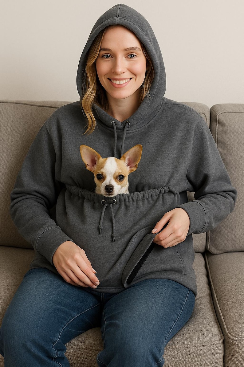 May include: A dark gray hoodie with a kangaroo pouch, featuring a small tan and white dog. The hoodie has a drawstring hood and is worn with blue jeans. The dog is looking directly at the camera.