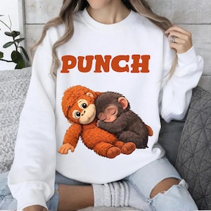 Op de afbeelding: Witte sweatshirt met het woord "PUNCH" in oranje letters. Daaronder een illustratie van twee cartoonapen die elkaar omarmen. De ene aap is oranje, de andere bruin.