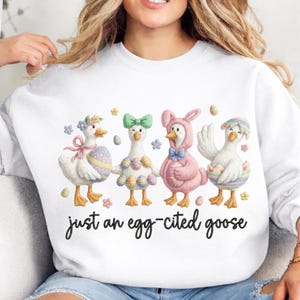 May include: White sweatshirt featuring four cartoon geese dressed in Easter-themed outfits. The geese are decorated with bows, egg-shaped designs, and a bunny costume. The text "just an egg-cited goose" is written in a script font.