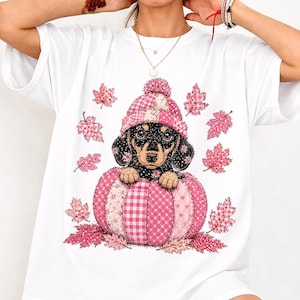 Könnte beinhalten: Ein weißes T-Shirt mit einer Grafik eines schwarz-braunen Dackelwelpen, der eine rosa karierte Strickmütze trägt. Der Welpe sitzt in einem großen rosa karierten Kürbis, umgeben von verstreuten rosa Herbstblättern mit verschiedenen Mustern.
