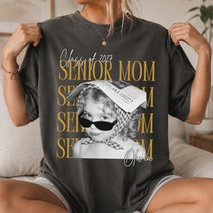 Könnte beinhalten: Ein anthrazitfarbenes Kurzarm-T-Shirt mit dem Text Class of 2027 SENIOR MOM in goldener Schrift. Das zentrale Motiv ist ein Schwarz-Weiß-Foto eines Kindes mit Sonnenbrille und Kopftuch, darunter steht der Name Olivia in Schreibschrift.