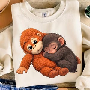 Könnte beinhalten: Cremefarbenes Sweatshirt mit einer Cartoon-Illustration von zwei sich umarmenden Affen. Ein Affe ist orange, der andere grau, beide mit geschlossenen Augen, was auf Schlaf hindeutet. Das Design ist mittig auf der Vorderseite des Kleidungsstücks platziert.