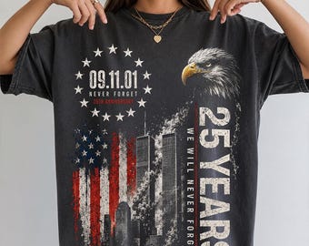 May include: A charcoal grey t-shirt featuring a graphic print with a bald eagle, the Twin Towers, and an American flag. Text includes 09.11.01, NEVER FORGET, 25TH ANNIVERSARY, and 25 YEARS WE WILL NEVER FORGET, surrounded by a circle of thirteen stars.