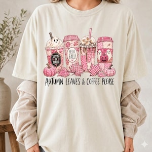 Könnte beinhalten: Ein cremefarbenes T-Shirt mit einer Grafik von vier rosafarbenen Kaffeebechern, die mit Geistern, Kürbissen und Herbstblättern verziert sind. Der Text lautet "Sippin on magic", "Hello, Fall!" und "Autumn leaves & coffee please" in Schwarz.