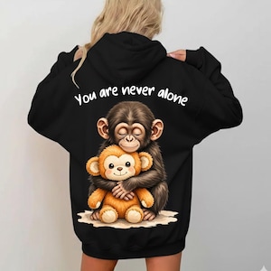 Könnte beinhalten: Schwarzer Kapuzenpullover mit einer Illustration von zwei sich umarmenden Zeichentrickaffen. Der weiße Text darüber lautet "You are never alone". Die Affen sind braun und orange.