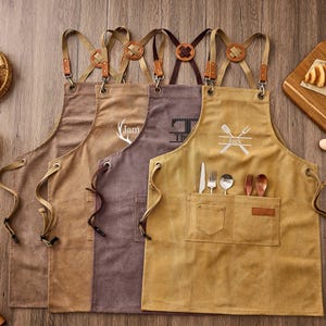 Puede incluir: Cuatro delantales de lona en colores beige, marrón y gris, cada uno con correas cruzadas con detalles en cuero, herrajes metálicos y un bolsillo frontal. Los delantales muestran varios diseños bordados, incluyendo el nombre Jack y cubiertos.