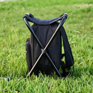 May include: Black backpack with an integrated folding stool. The backpack has a black frame and seat. The backpack is on green grass.