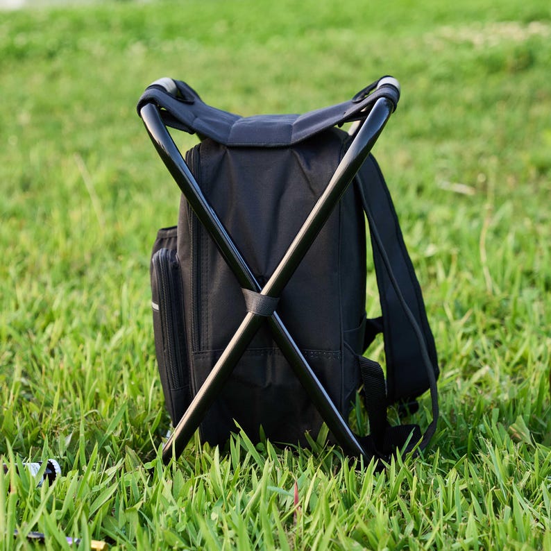May include: Black backpack with an integrated folding stool. The backpack has a black frame and seat. The backpack is on green grass.