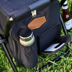 May include: Black picnic cooler with a built-in chair. The cooler has a front zipped pocket, a side pocket for a bottle, and a brown leather patch. A bottle of beer is visible in the background.