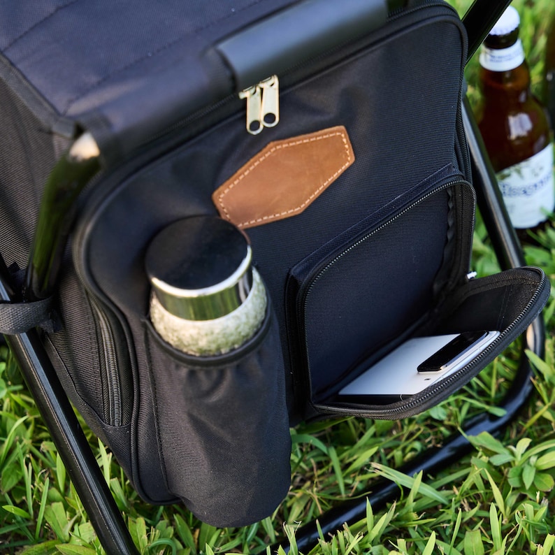May include: Black picnic cooler with a built-in chair. The cooler has a front zipped pocket, a side pocket for a bottle, and a brown leather patch. A bottle of beer is visible in the background.