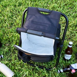 May include: A black folding chair with an integrated cooler, positioned on grass. The cooler is open, displaying a light grey interior. Several beer bottles are scattered around the chair.