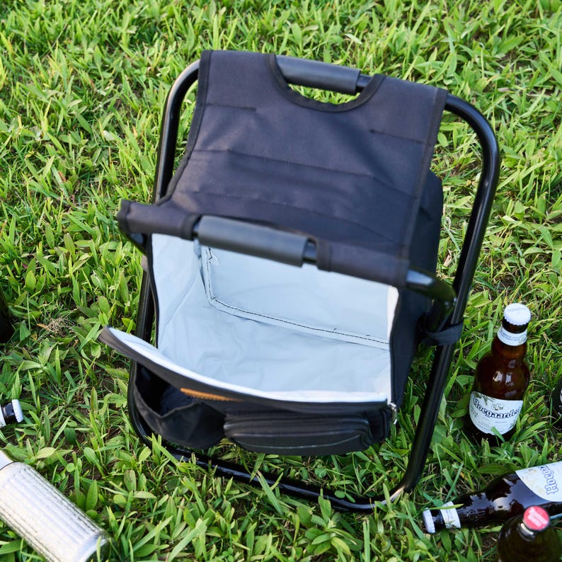 May include: A black folding chair with an integrated cooler, positioned on grass. The cooler is open, displaying a light grey interior. Several beer bottles are scattered around the chair.