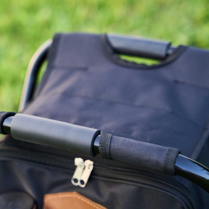 May include: Close-up of a black rolling cart with a padded handle and a black fabric bag. The handle is black with a grey padded grip. The bag has a brown leather patch and a double zip.