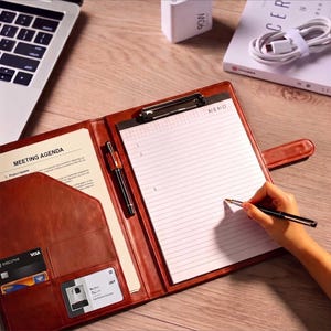 May include: A brown leather portfolio folder featuring a metal clipboard, a lined memo pad with the text MEMO, a pen holder with a black pen, and interior pockets containing a meeting agenda document, a credit card, and an identification card.