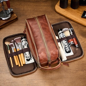 May include: A brown leather wash bag with a zip, open to show makeup brushes, shaving supplies, and tubes of cream. The bag is on a wooden surface, with a handle on the side.