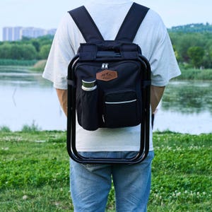 May include: Black backpack with an integrated folding chair. The backpack has a brown leather patch with a mountain logo and the word "James". It features a side pocket for a water bottle and a front pocket with a white stripe. The chair frame is black.