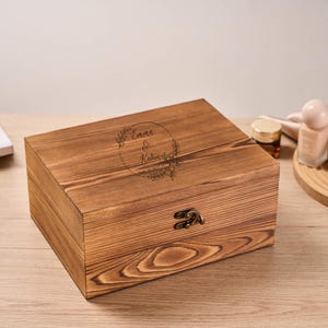 Könnte beinhalten: Eine rechteckige Holzkiste mit Scharnierdeckel und Metallverschluss. Die Kiste besteht aus hellbraunem Holz mit sichtbarer Maserung. Der Deckel zeigt ein kreisförmiges Blumendesign mit den Namen "Emma & Kevin" und dem Datum "10. Juni 2023".