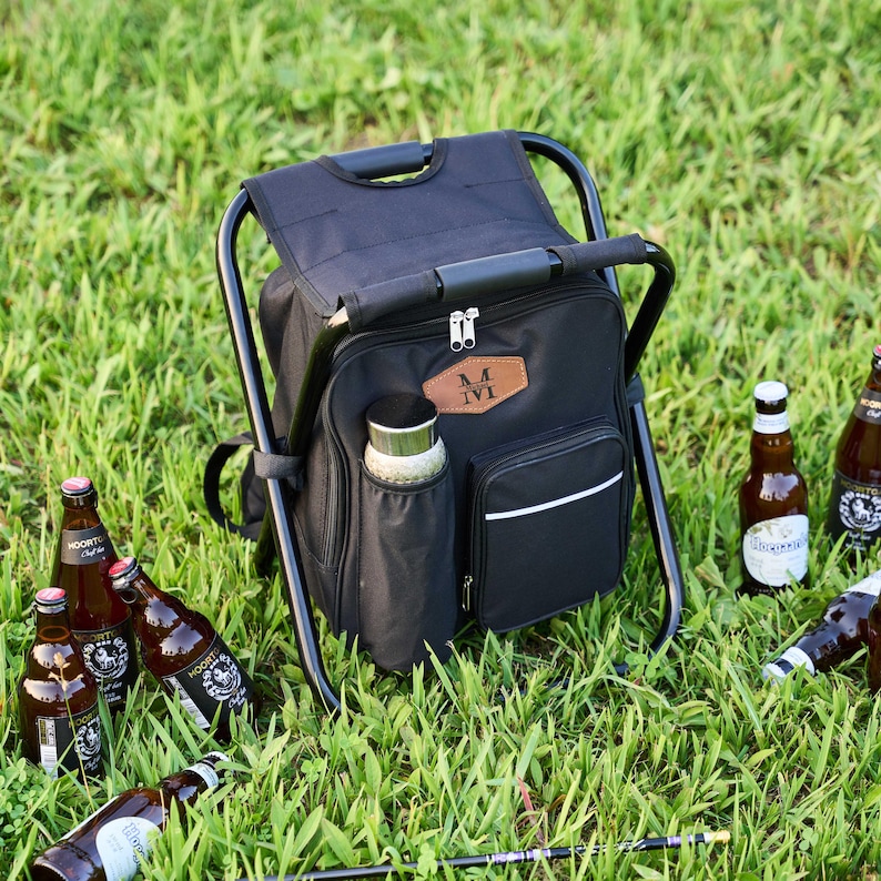 May include: Black cooler chair with a black frame, a handle, and a side pocket. The cooler has a brown leather patch with the initials "M IVI". Several beer bottles are scattered on the green grass.