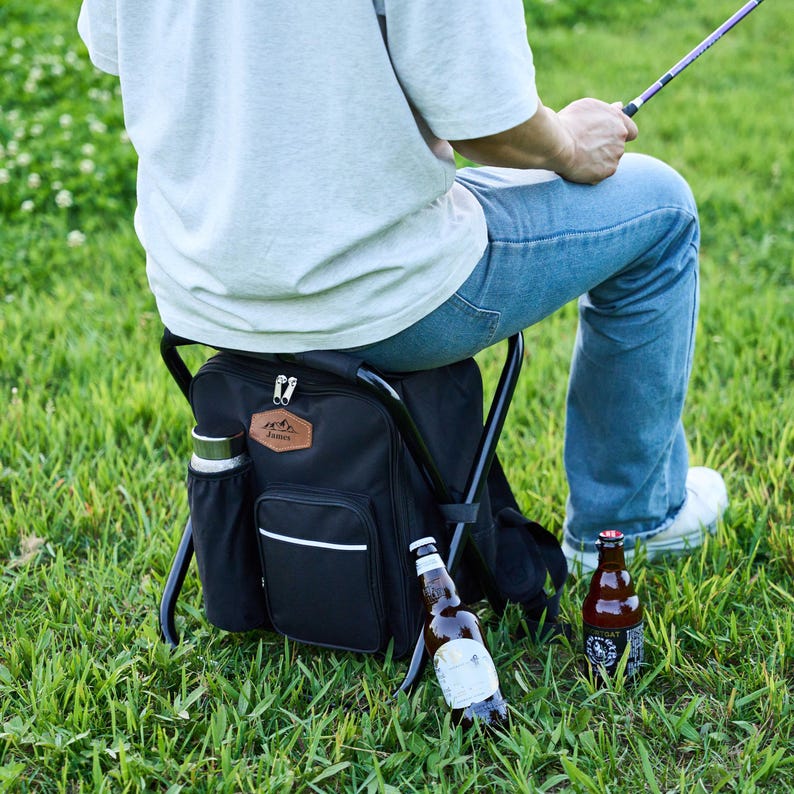May include: A black backpack chair with a brown leather patch and a side pocket holding a green insulated bottle. Two beer bottles are on the grass. The person is wearing blue jeans and white trainers.