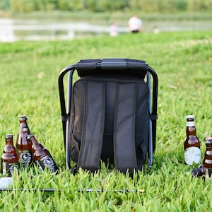 May include: A black backpack chair with a metal frame is set on grass. Several brown beer bottles are scattered around the chair. The background shows a lake and people.