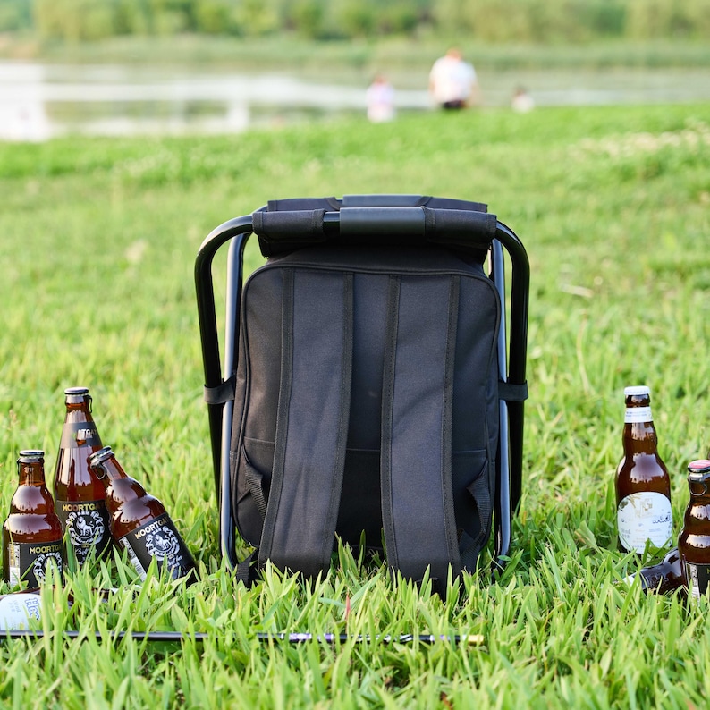 May include: A black backpack chair with a metal frame is set on grass. Several brown beer bottles are scattered around the chair. The background shows a lake and people.