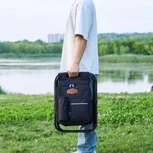 May include: A black backpack chair with a brown leather patch and white trim is held by a person. The chair has a black metal frame and a side pocket. The background shows a lake and green grass.