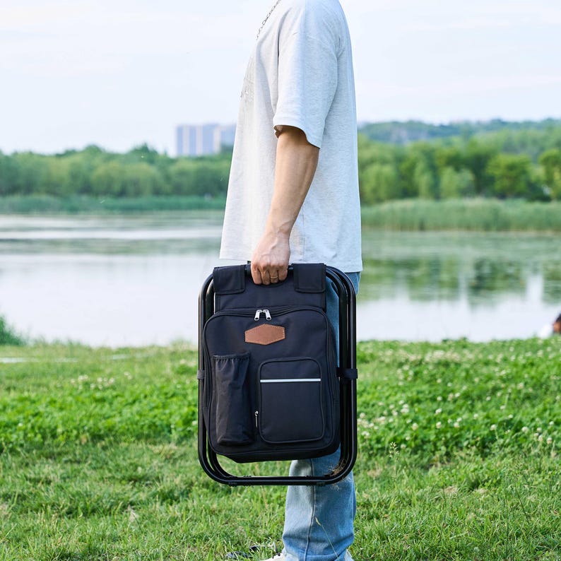 May include: A black backpack chair with a brown leather patch and white trim is held by a person. The chair has a black metal frame and a side pocket. The background shows a lake and green grass.
