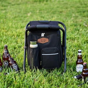 May include: A black cooler backpack with an integrated folding chair, positioned on grass. The backpack features a side pocket for a water bottle and a front pocket with a white stripe. Several beer bottles are scattered around the cooler.
