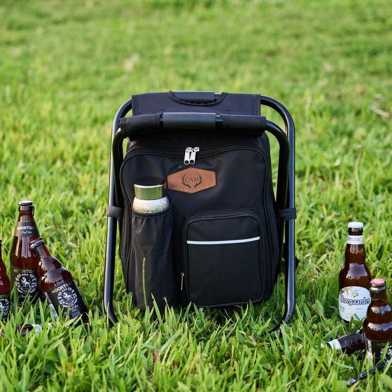 May include: A black cooler backpack with an integrated folding chair, positioned on grass. The backpack features a side pocket for a water bottle and a front pocket with a white stripe. Several beer bottles are scattered around the cooler.