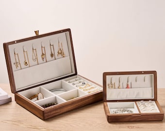 May include: Two open wooden jewelry boxes with a light interior. The larger box displays necklaces, rings, and a watch. The smaller box holds rings and earrings. A gold necklace lies on the wooden surface.