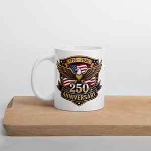 May include: White ceramic mug featuring a patriotic design. The design includes a bald eagle with outstretched wings, the American flag, and the text "1776-2026 250 Anniversary". The mug is placed on a wooden surface.