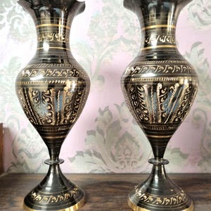 Op de afbeelding: Twee sierlijke zwarte vazen met gouden accenten. De vazen hebben een uitlopende, gerimpelde bovenkant en een slanke hals die uitloopt in een bolvormig lichaam. Sierlijke gouden patronen sieren het lichaam en de basis is ook goudkleurig. De vazen staan op een houten oppervlak.