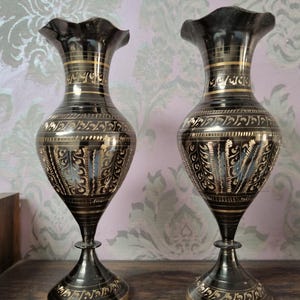 Può includere: Due vasi in metallo nero con accenti dorati e disegni intricati. I vasi hanno un'ampia apertura e una base stretta.