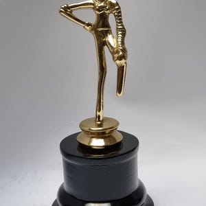 May include: A gold-colored metal trophy featuring a female figure in a dancing pose. The figure is standing on a black wooden base.