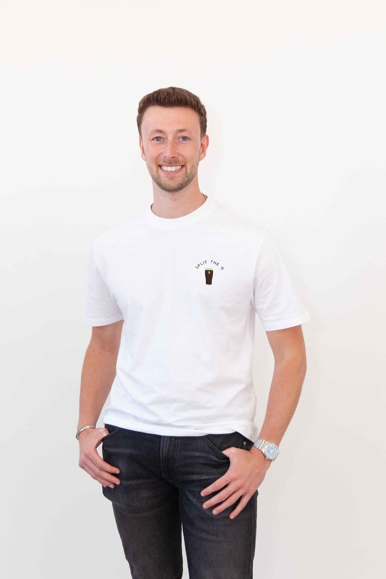 May include: A man wearing a white t-shirt with the text "Split the G" and a graphic of a pint glass. He is wearing black jeans and a silver watch.