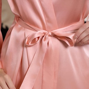 May include: A close-up of a silky, blush-colored robe tied with a matching sash. The fabric has a subtle sheen, and the bow is neatly knotted. The robe's design features a wrap-around style, suggesting comfort and elegance.
