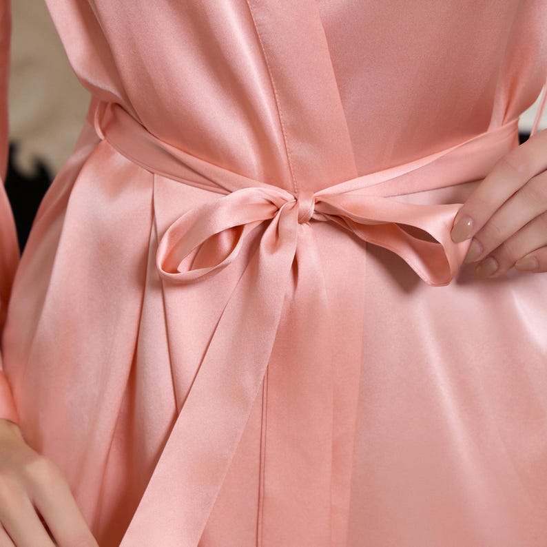 May include: A close-up of a silky, blush-colored robe tied with a matching sash. The fabric has a subtle sheen, and the bow is neatly knotted. The robe's design features a wrap-around style, suggesting comfort and elegance.