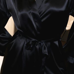 May include: A black satin robe with a wrap-around design and a matching tie belt. The robe features wide sleeves and a smooth, shiny texture. The tie is knotted at the waist, creating a flattering silhouette. The robe is a luxurious piece of sleepwear.