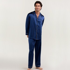 Op de afbeelding: Een donkerblauwe satijnen pyjamaset met witte piping. Het bovenstuk heeft een knoopsluiting en een kraag. De broek heeft een rechte pijp.