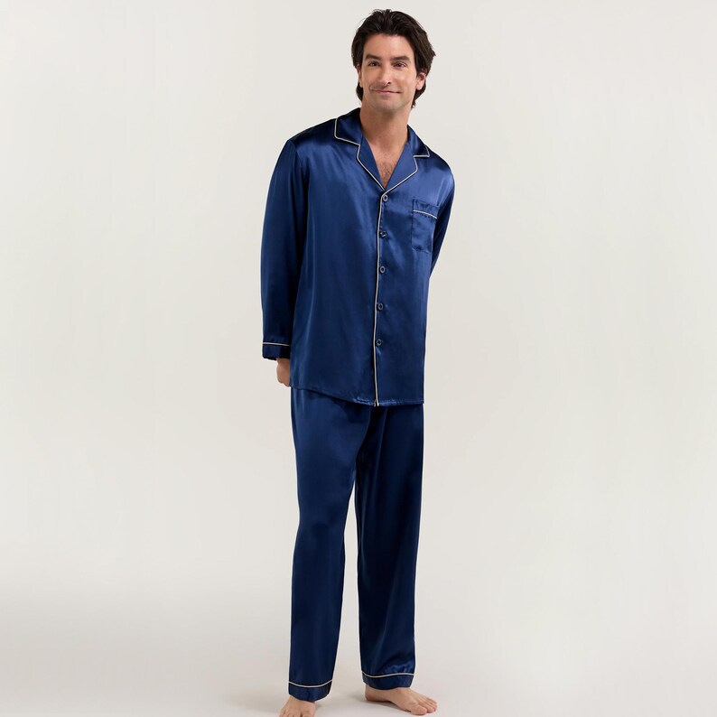 Op de afbeelding: Een donkerblauwe satijnen pyjamaset met witte piping. Het bovenstuk heeft een knoopsluiting en een kraag. De broek heeft een rechte pijp.