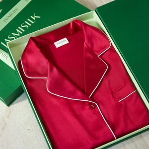 Op de afbeelding: Een rode zijden pyjama top met witte piping rond de kraag en manchetten. De top heeft een knoopsluiting en een zakje aan de linkerkant. De top zit in een cadeaudoos met een groen deksel waarop "JASMISILK" staat.