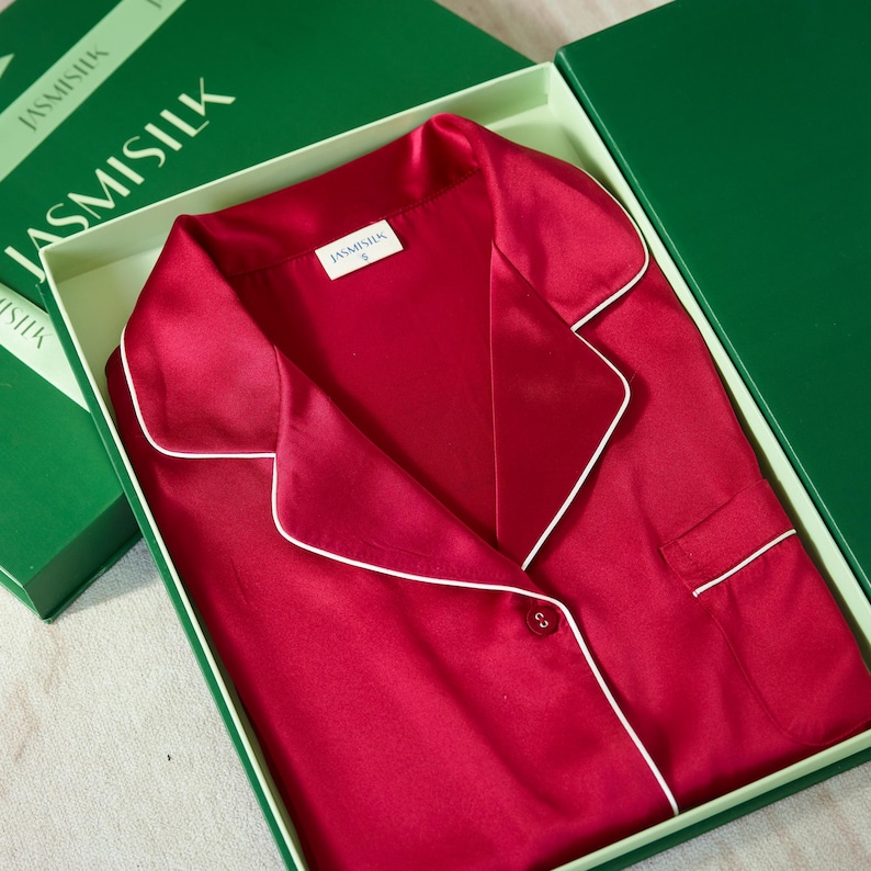 Op de afbeelding: Een rode zijden pyjama top met witte piping rond de kraag en manchetten. De top heeft een knoopsluiting en een zakje aan de linkerkant. De top zit in een cadeaudoos met een groen deksel waarop "JASMISILK" staat.
