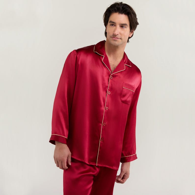 Op de afbeelding: Een lang mouwig rood zijden pyjama top met een witte bies. De top heeft een knoopsluiting aan de voorkant en een zak aan de linkerkant.